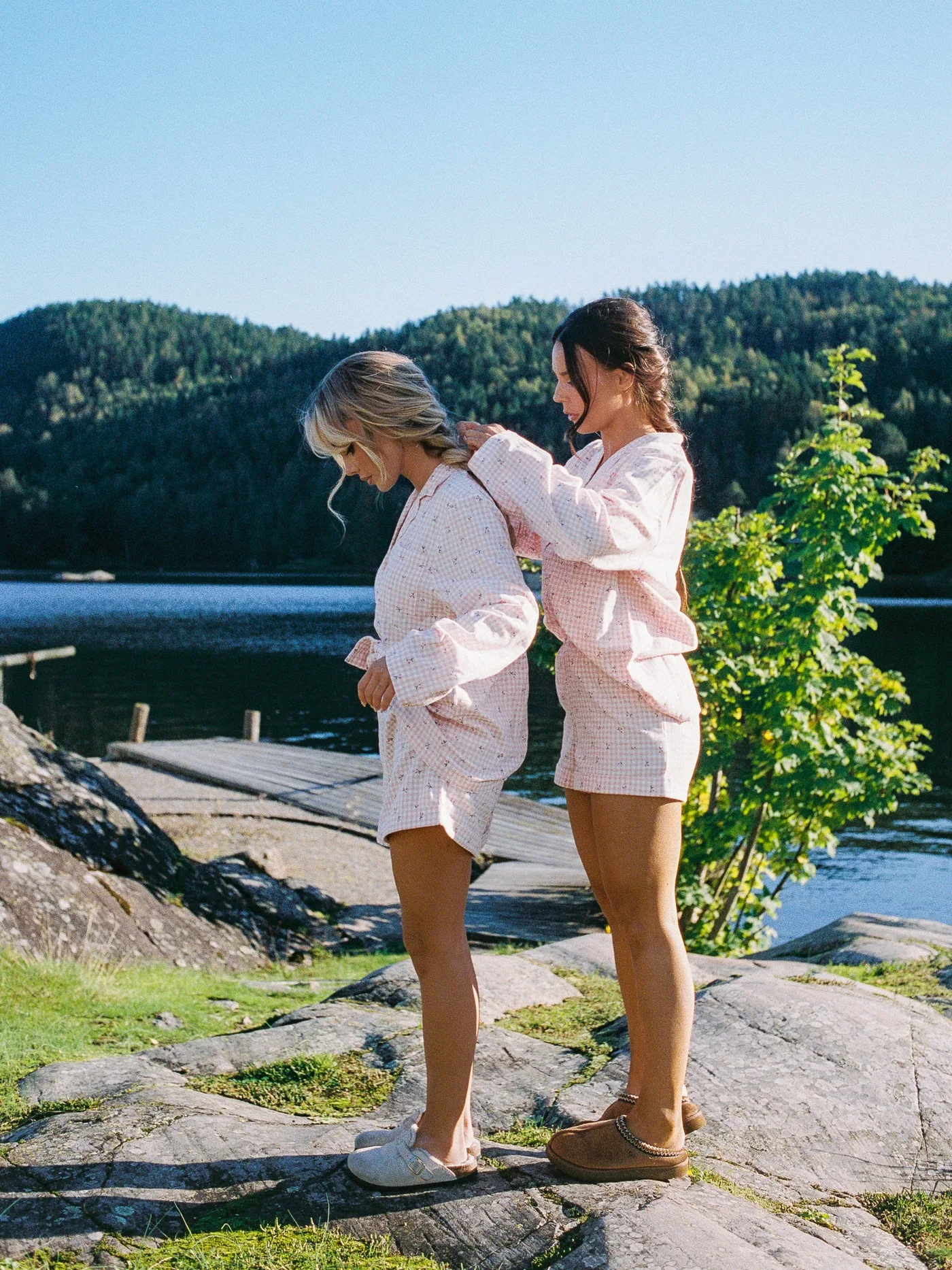 To modeller i rosa rutete pysj ved vannet, den ene fletter håret til den andre